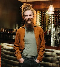 a bearded man standing in front of a bar full of wine bottles